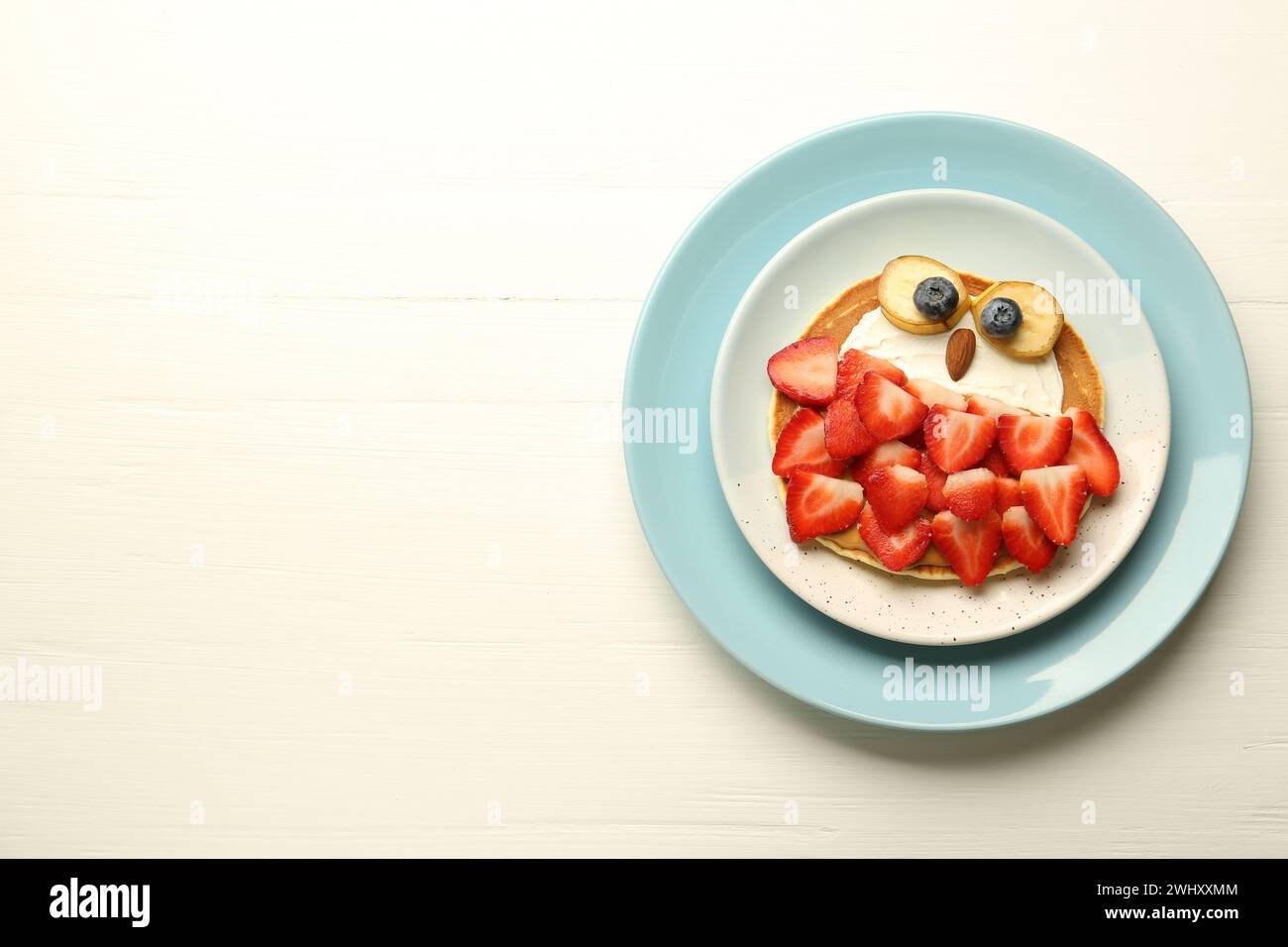 Kreatives Servieren für Kinder. Teller mit süßer Eule aus Pfannkuchen, Erdbeeren, Creme, Banane und Mandel auf weißem Holztisch, Blick von oben. Raum für Tex Stockfoto