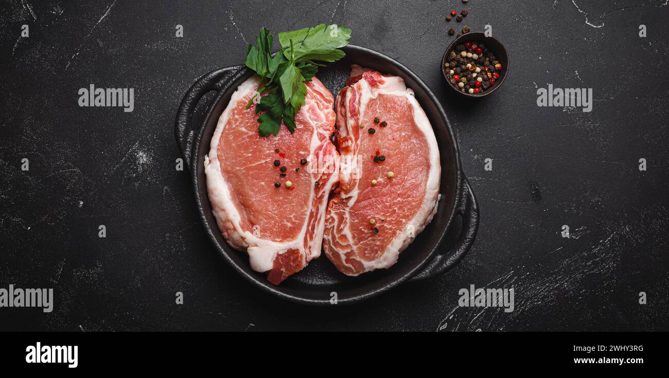 Schneiden Sie rohe Schweinefleisch-Steaks mit Gewürzen in einer schwarzen gusseisernen Pfanne auf dunklem rustikalem Steinhintergrund von oben, bereit zum Rösten. Po Stockfoto