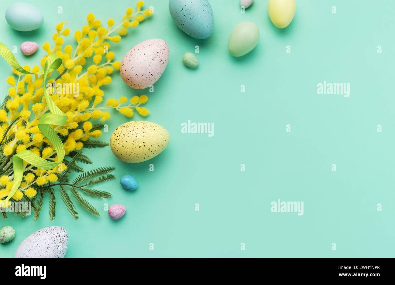 Ein kreatives Arrangement aus pastellfarbenen Ostereiern, Mimosa-Zweigen und Bonbons auf einer weichen grünen Oberfläche, das die Frühlingsfeier symbolisiert Stockfoto