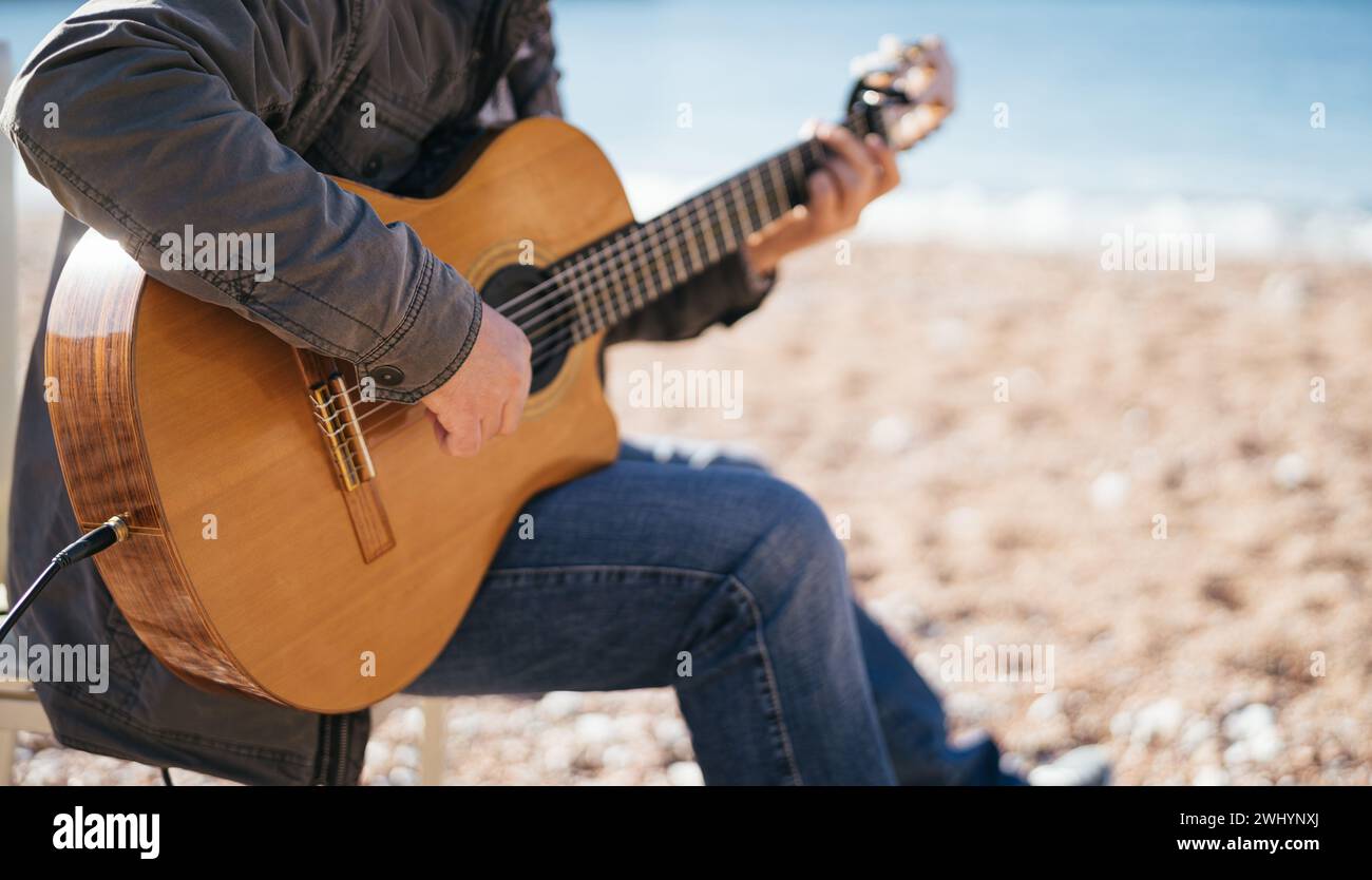 Der Musiker spielt Akustikgitarre, während er auf einem Stuhl am Strand sitzt. Abgeschnitten. Gesichtslos Stockfoto