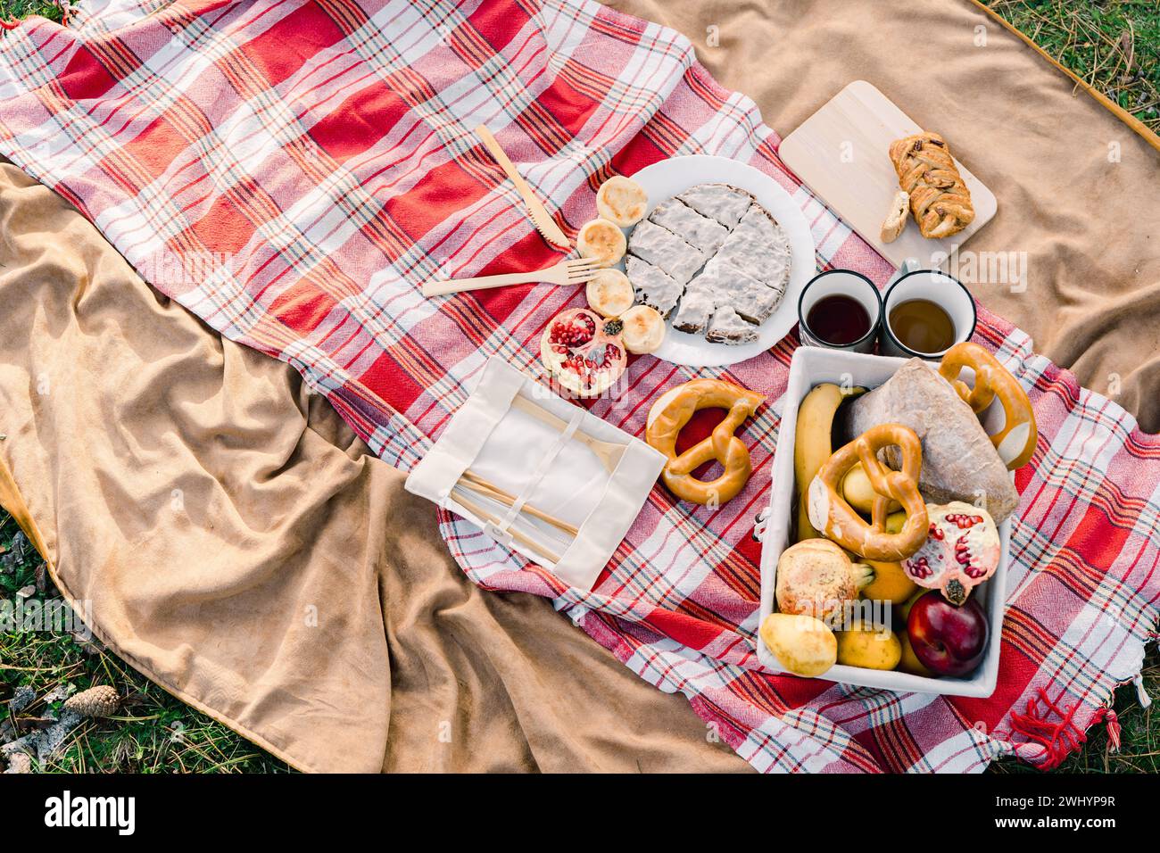 Ein Korb mit Gebäck und Obst steht neben Kaffeetassen auf einer Decke auf grünem Gras Stockfoto