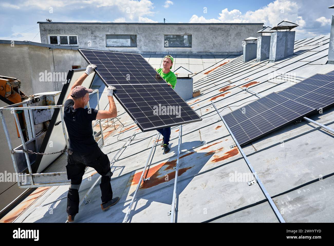 Arbeiter, die Solarpaneele auf dem Metalldach des Hauses mit Hilfe eines Kranaufzugs bauen. Zwei Monteure tragen Photovoltaik-Solarmodul im Freien. Konzept der Erzeugung erneuerbarer Energien. Stockfoto