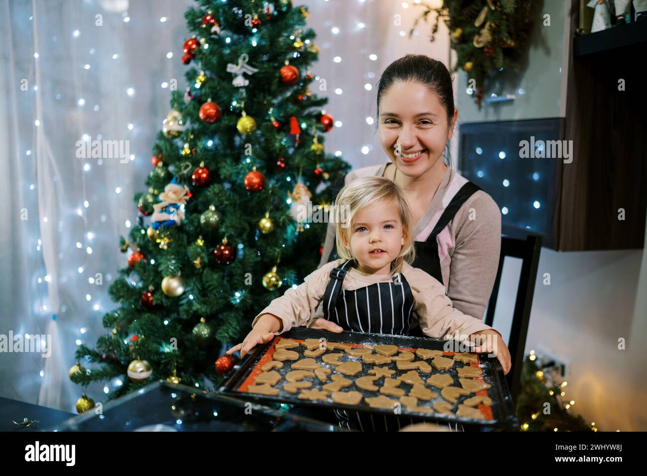 Lächelnde Mutter mit einer kleinen Tochter, die am Tisch sitzt und ein Blatt roher Weihnachtsplätzchen hat Stockfoto