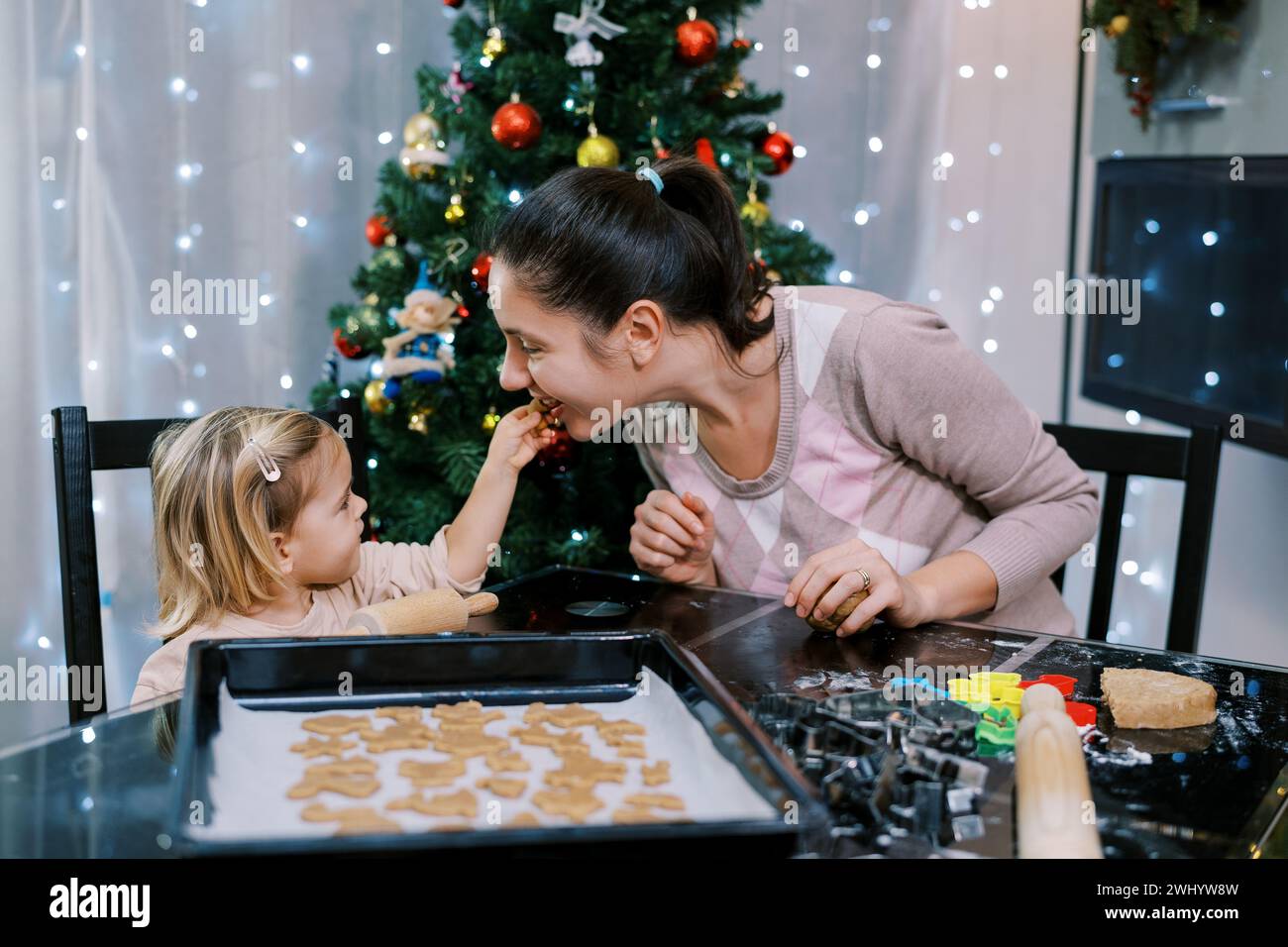 Die kleine Tochter verwöhnt ihre Mutter mit einem Keks, während sie an einem Tisch mit rohen Keksen auf einem Tablett sitzt Stockfoto