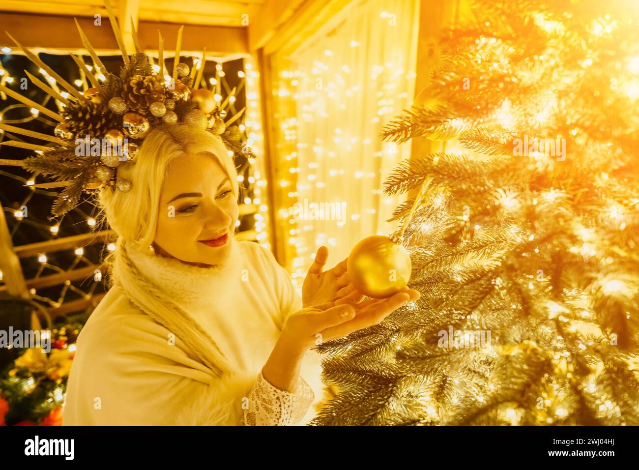 Eine blonde Frau in weißem Kleid und eine Krone aus goldenen Ornamenten schmücken den Weihnachtsbaum mit goldenen Ornamenten und Lichtern. Der Baum ist mit Gold verziert Stockfoto