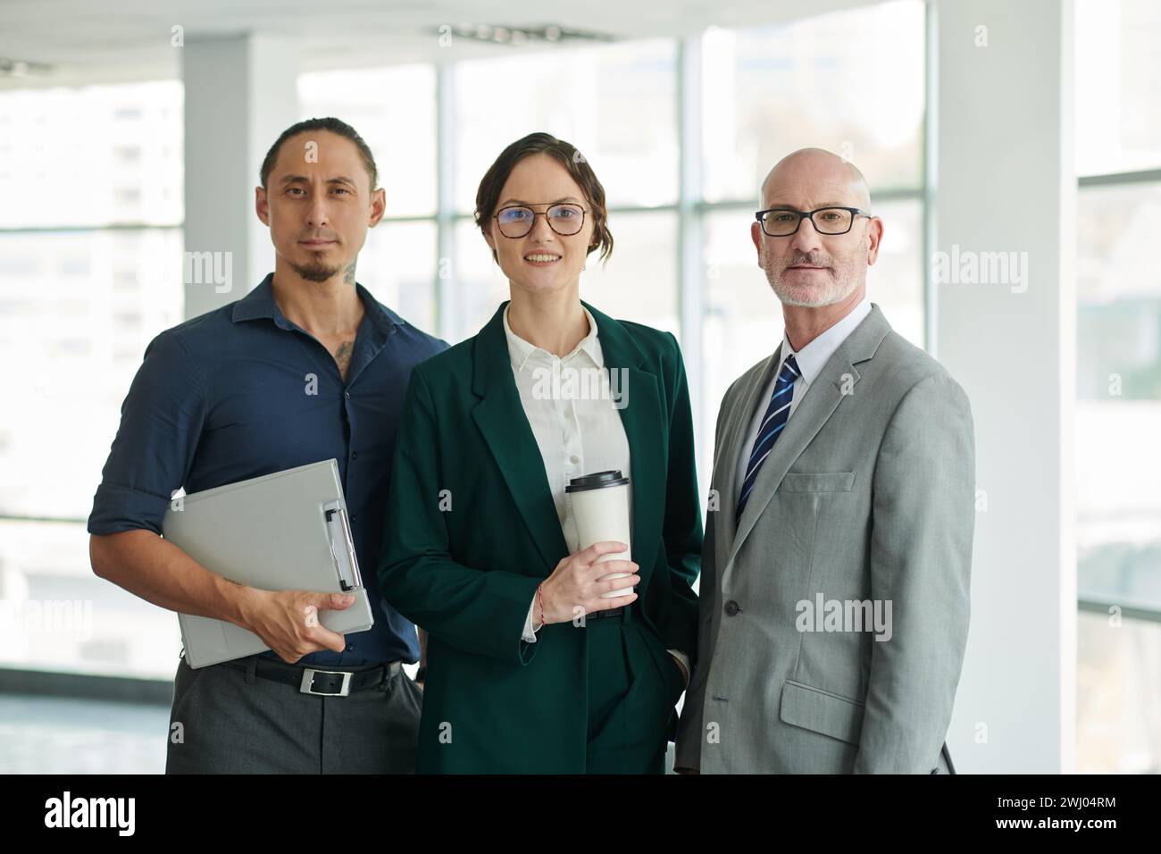 Selbstbewusster Direktor des Unternehmens in grauem Anzug neben jungen interkulturellen Mitarbeitern, während jeder in die Kamera schaut Stockfoto