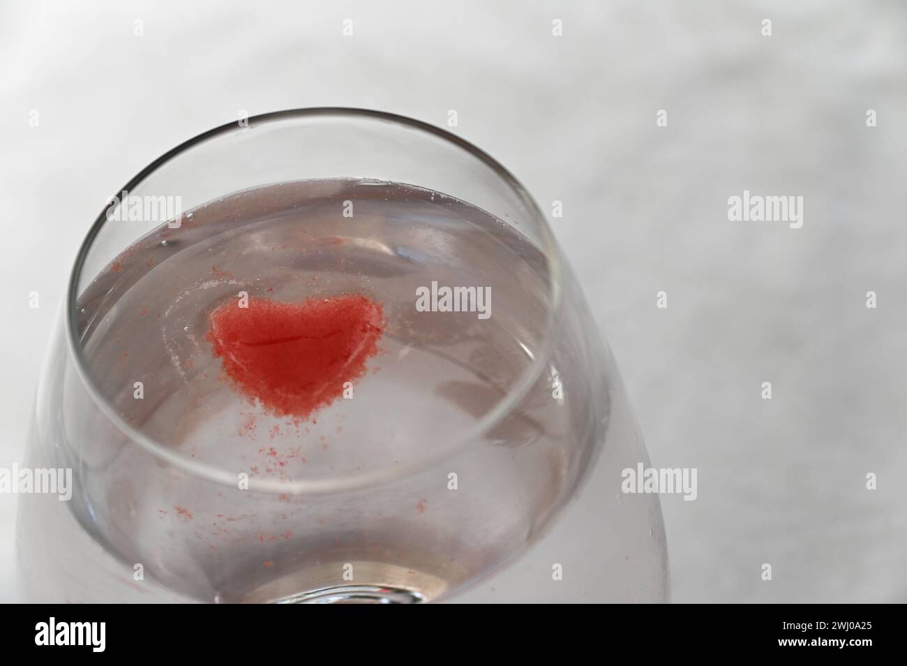 Ein Glas ist voller klarer Flüssigkeit und ein leuchtend roter, herzförmiger Eiswürfel, der darin schwimmt Stockfoto