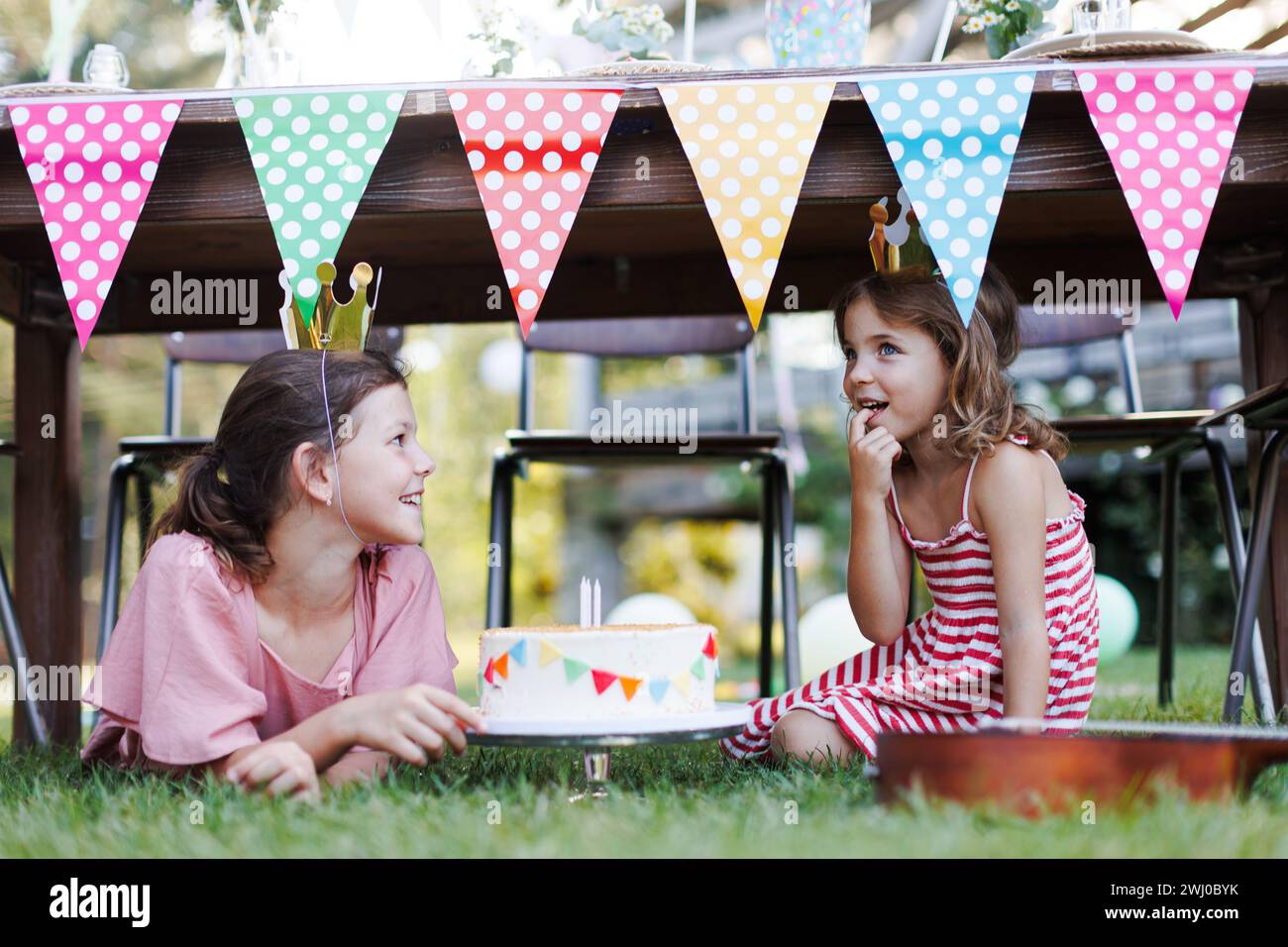 Süße Mädchen mit Papierkronen, die Geburtstagskuchen unter dem Tisch essen. Geburtstagsgartenparty für Kinder. Stockfoto