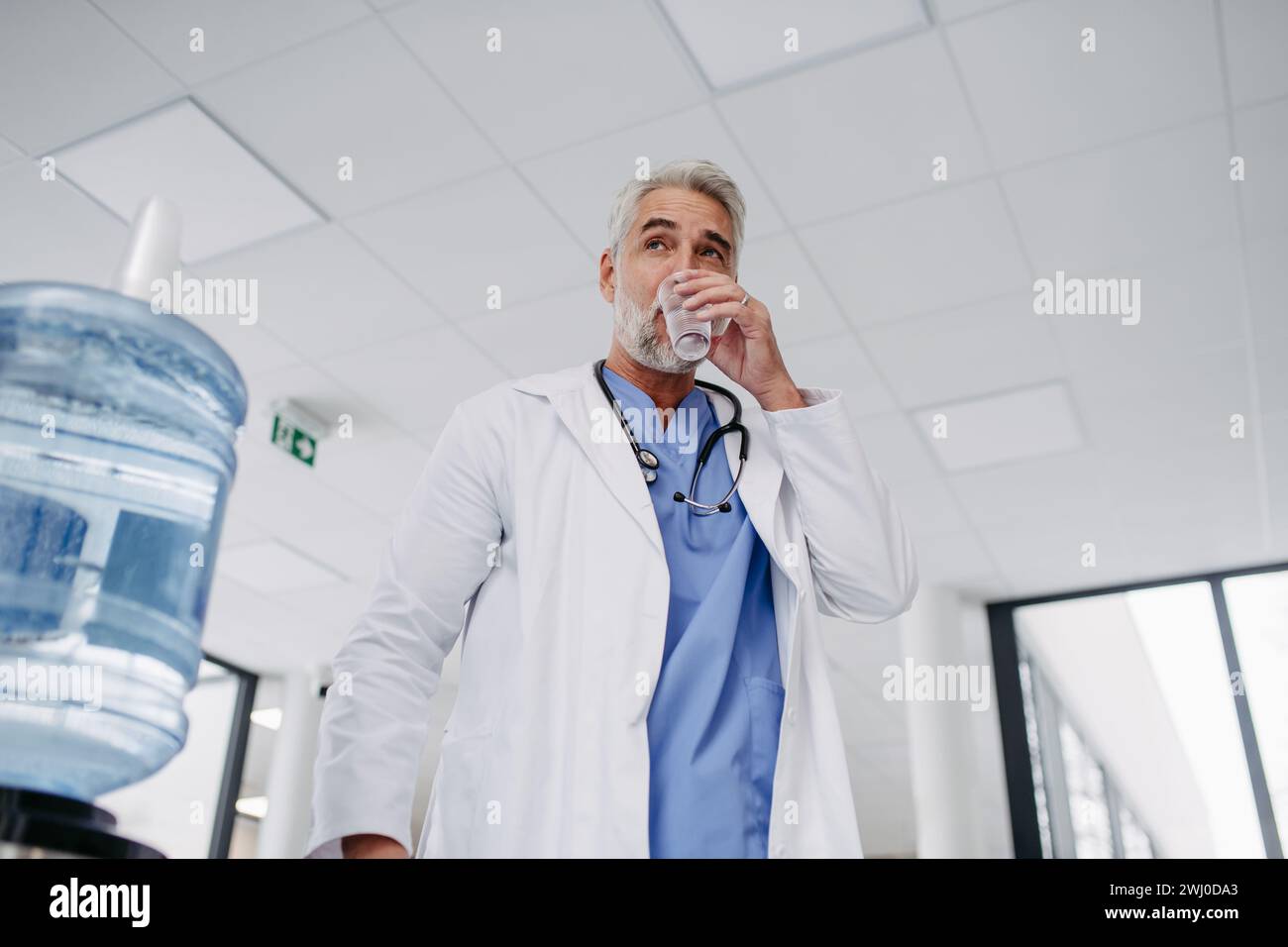 Gutaussehender Arzt, der eine Pause während der Arbeitsschicht im Krankenhaus macht, Trinkwasser aus dem Wasserspender in der Krankenhauslobby trinkt. Stockfoto