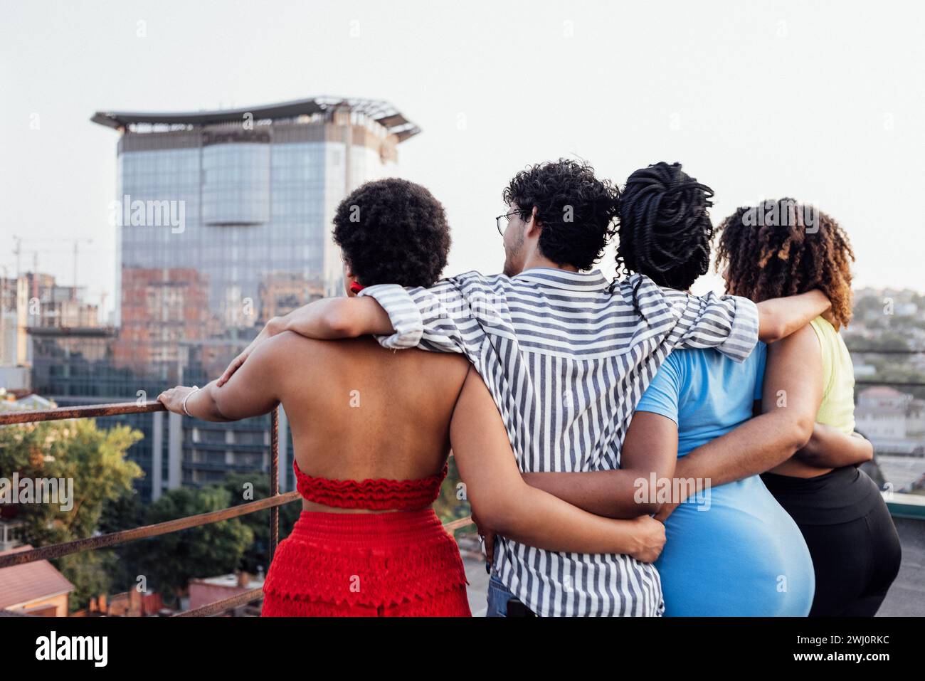 Vier Freunde umarmen sich und stehen mit dem Rücken auf dem Dach des Hauses. Mann und afrikanische Frauen in lässiger Kleidung schauen sich die Stadt an Stockfoto