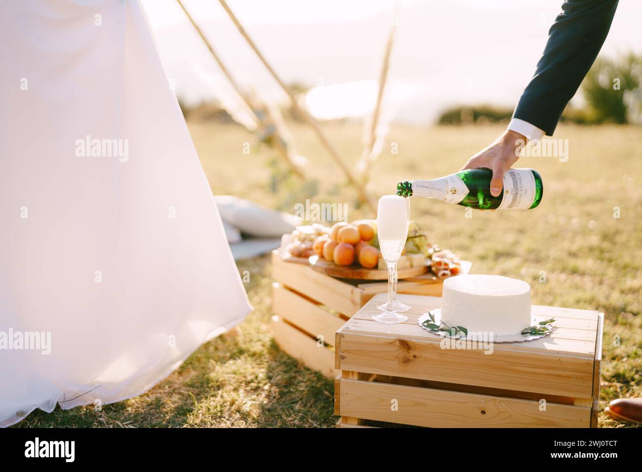 Der Bräutigam gießt Champagner in Gläser auf einer Holzkiste mit einem Kuchen neben der Braut. Abgeschnitten Stockfoto