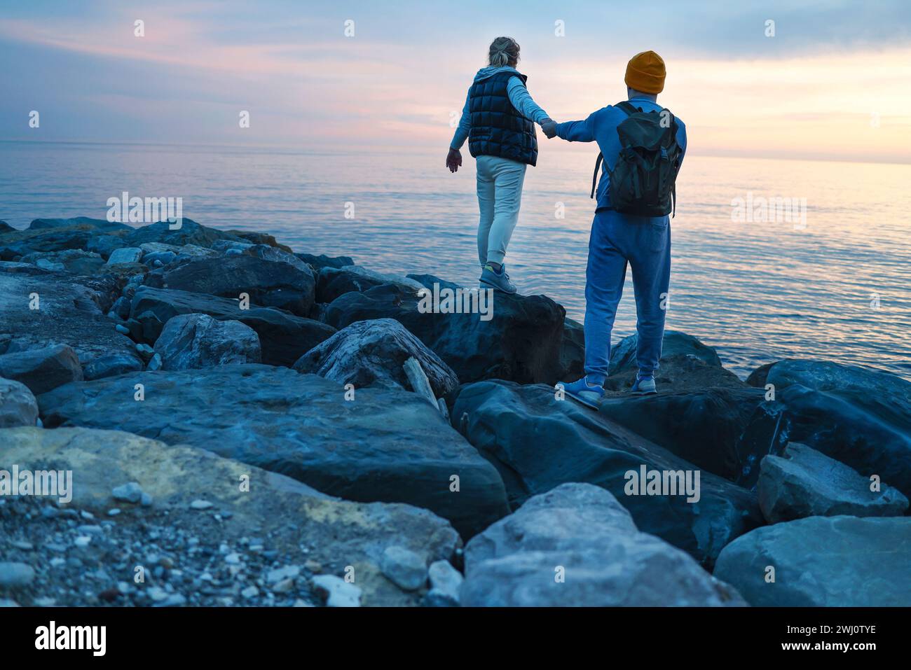 Ein junges Paar spaziert bei Sonnenuntergang am Meer entlang. Ein Mann und eine Frau in lässiger Kleidung stehen zurück. Stockfoto