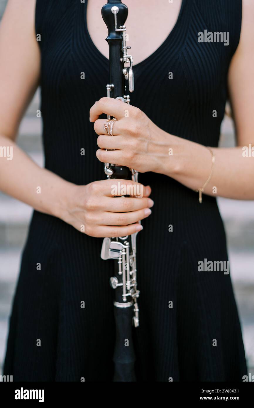Das Mädchen des Musikers in einem schwarzen Kleid hält eine Klarinette in den Händen vor der Brust. Abgeschnitten. Gesichtslos Stockfoto