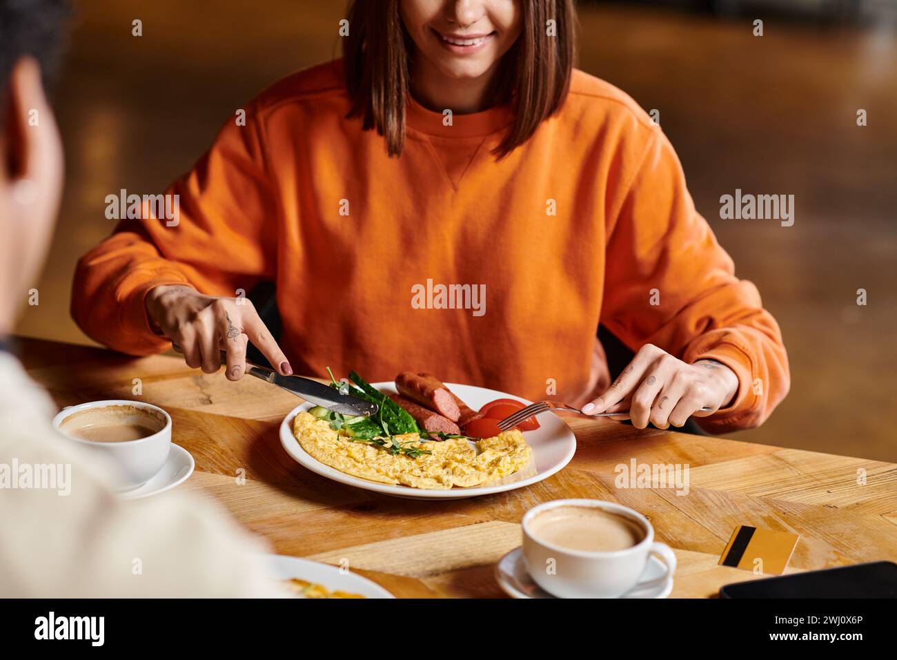 Glückliche Frau, die in der Nähe ihrer Tasse Kaffee frühstückt, während sie in der Nähe des schwarzen Freundes leckeres Essen genießt Stockfoto