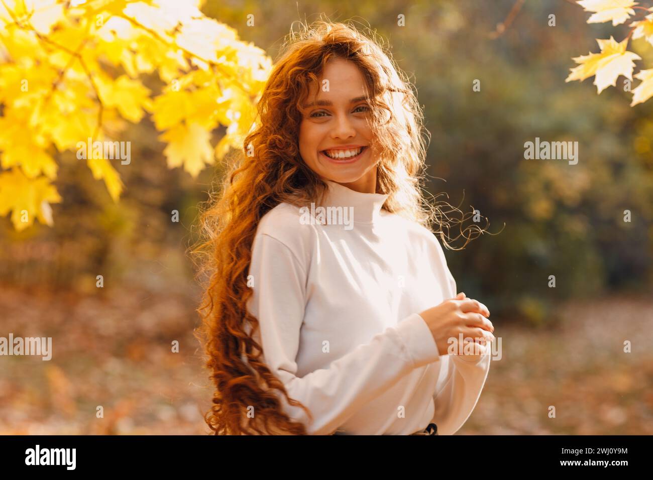 Lächelnde junge Frau genießt das Herbstwetter im Park mit den gelben Blättern an den Ästen. Stockfoto