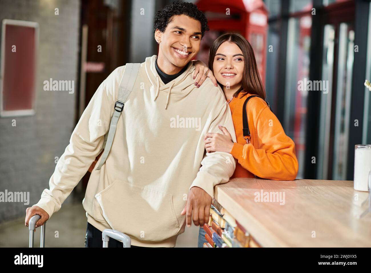 Fröhliches, interrassisches Paar lächelt an der Rezeption des Hostels, scheinbar aufgeregt von ihrer Reise Stockfoto