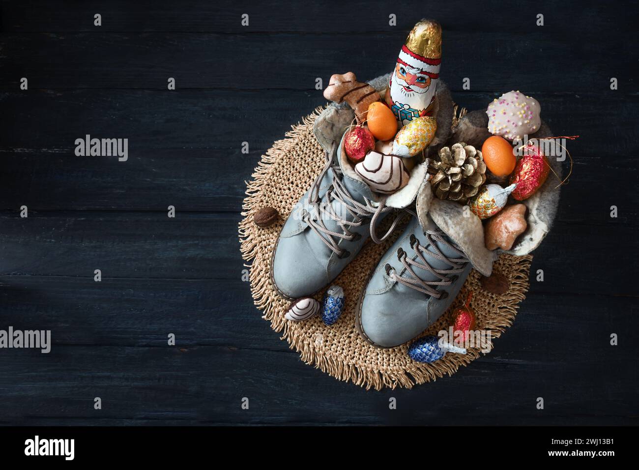 Winterstiefel gefüllt mit einem Schokoladen-Weihnachtsmann, Leckereien und Weihnachtsdekoration auf dunkelblauem Holz für Nikolaus Day oder German Nikolau Stockfoto