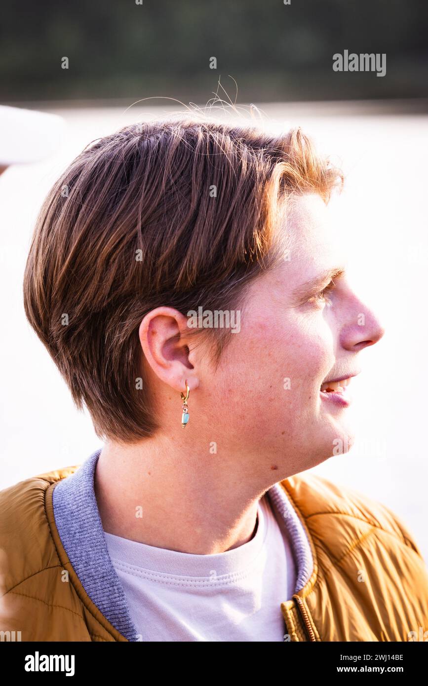 Dieses Bild zeigt ein Seitenprofil eines androgynen Individuums vor einem sanft verschwommenen natürlichen Hintergrund. Das Motiv scheint mitten im Lächeln zu sein, was dem Porträt ein Gefühl von Wärme und Positivität verleiht. Sie haben ein kurzes, ordentlich gestyltes Haar und tragen einen einfachen Ohrring, ein weißes T-Shirt und eine gelbe Jacke mit blauem Kragen, was einen lässigen und bequemen Stil andeutet. Die Beleuchtung ist weich und warm, was wahrscheinlich auf einen Untergang oder Sonnenaufgang hinweist, der den Eigenschaften ein sanftes Leuchten verleiht. Dieses Porträt fängt einen Moment ein, der sich sowohl offen als auch intim anfühlt, und hebt die r des Einzelnen hervor Stockfoto
