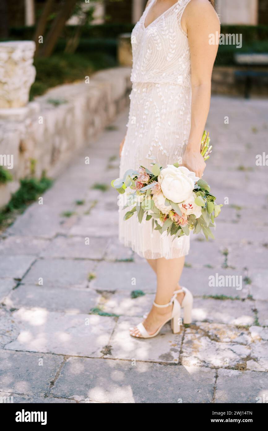 Braut mit einem Blumenstrauß steht halb gedreht auf den Pflastersteinen. Abgeschnitten. Gesichtslos Stockfoto