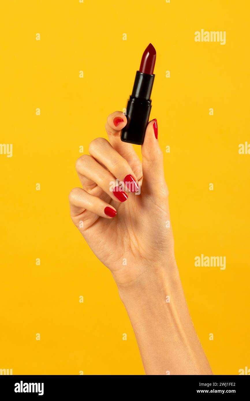 Anonyme schlanke junge weibliche Hand mit offenem Lippenstift im schlanken Zeigefinger und Daumenfinger mit nagellack in Licht vor leuchtendem gelbem Hintergrund Stockfoto