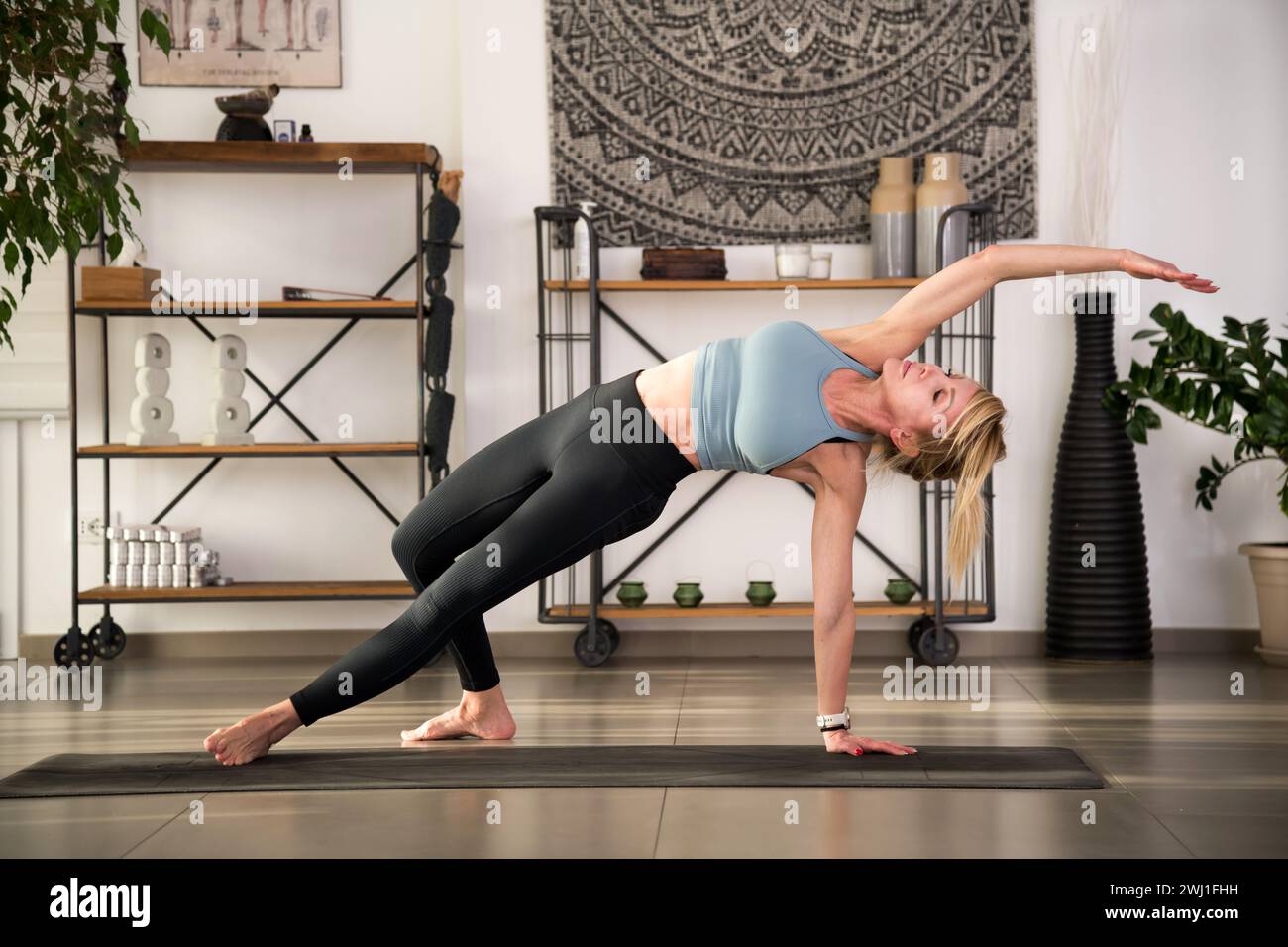 Flexible barfuß-Dame in Aktivkleidung, die Camatkarasana in Innenräumen zur Entspannung während der Yoga-Session an Regalen mit Topfpflanzen und Wandteppichen übt Stockfoto