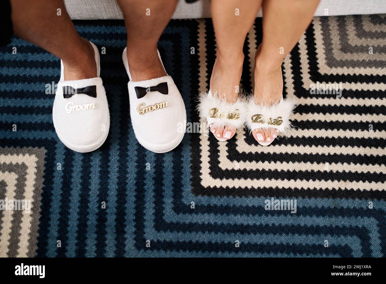 Männliche und weibliche Füße in Hausschuhen stehen auf einem gestreiften Teppich. Bildunterschrift: Bräutigam. Braut. Abgeschnitten, ohne Gesicht Stockfoto