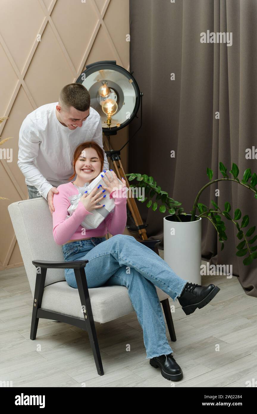 Ein Mann schenkt einem Mädchen ein Geschenk. Ein Mann umarmt ein Mädchen von hinten. Das Mädchen sitzt auf einem Stuhl. Stockfoto