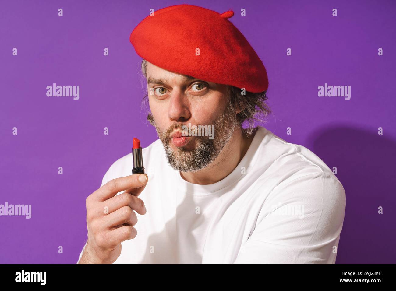 Lustiger Mann mittleren Alters mit roter Baskenmütze hält Lippenstift in der Hand vor violettem Hintergrund Stockfoto