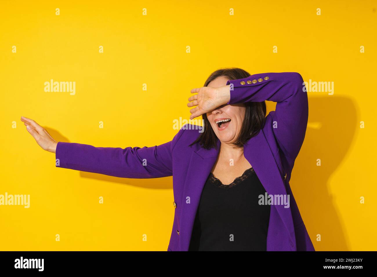 Fröhliche Frau mittleren Alters mit violettem Blazer, die vor gelbem Hintergrund tanzt Stockfoto