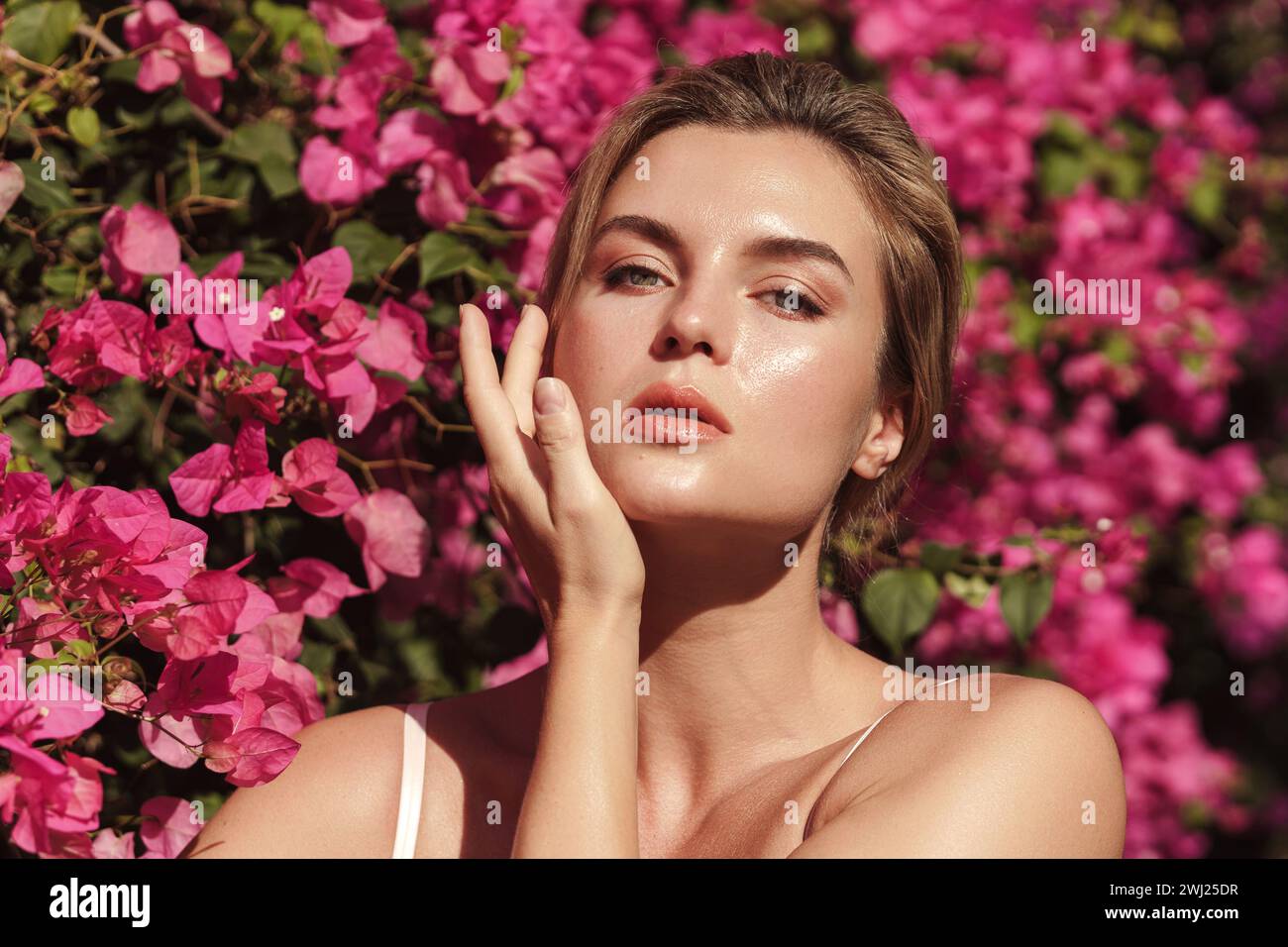 Schöne blonde Frau mit glatter Haut und natürlichem Make-up, während sie vor dem Hintergrund eines Busches mit zarten rosa Blüten posiert Stockfoto