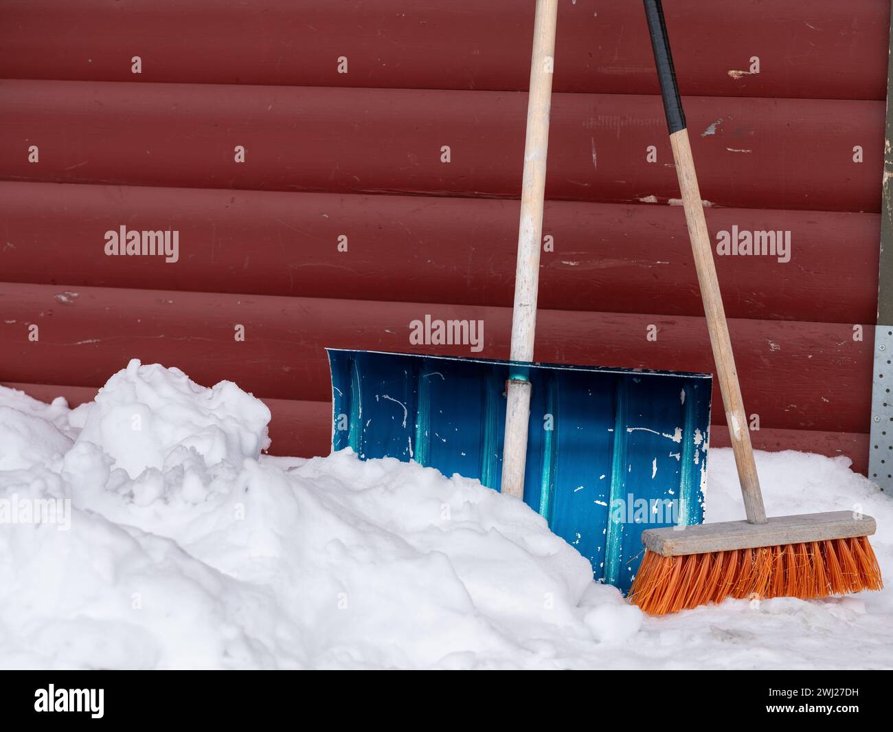 Shuffle und Besen stapelten sich an einer roten Wand mit Schnee auf dem Boden. Inspiration für die Arbeit im Winter Stockfoto