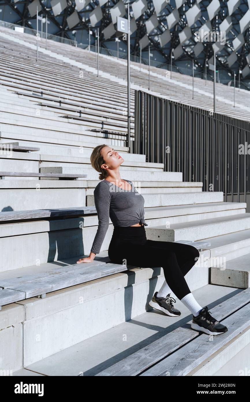 Sportlerin in weiblicher Sportbekleidung, die auf der Bank von Tribünen im Freiluftstadion sitzt Stockfoto