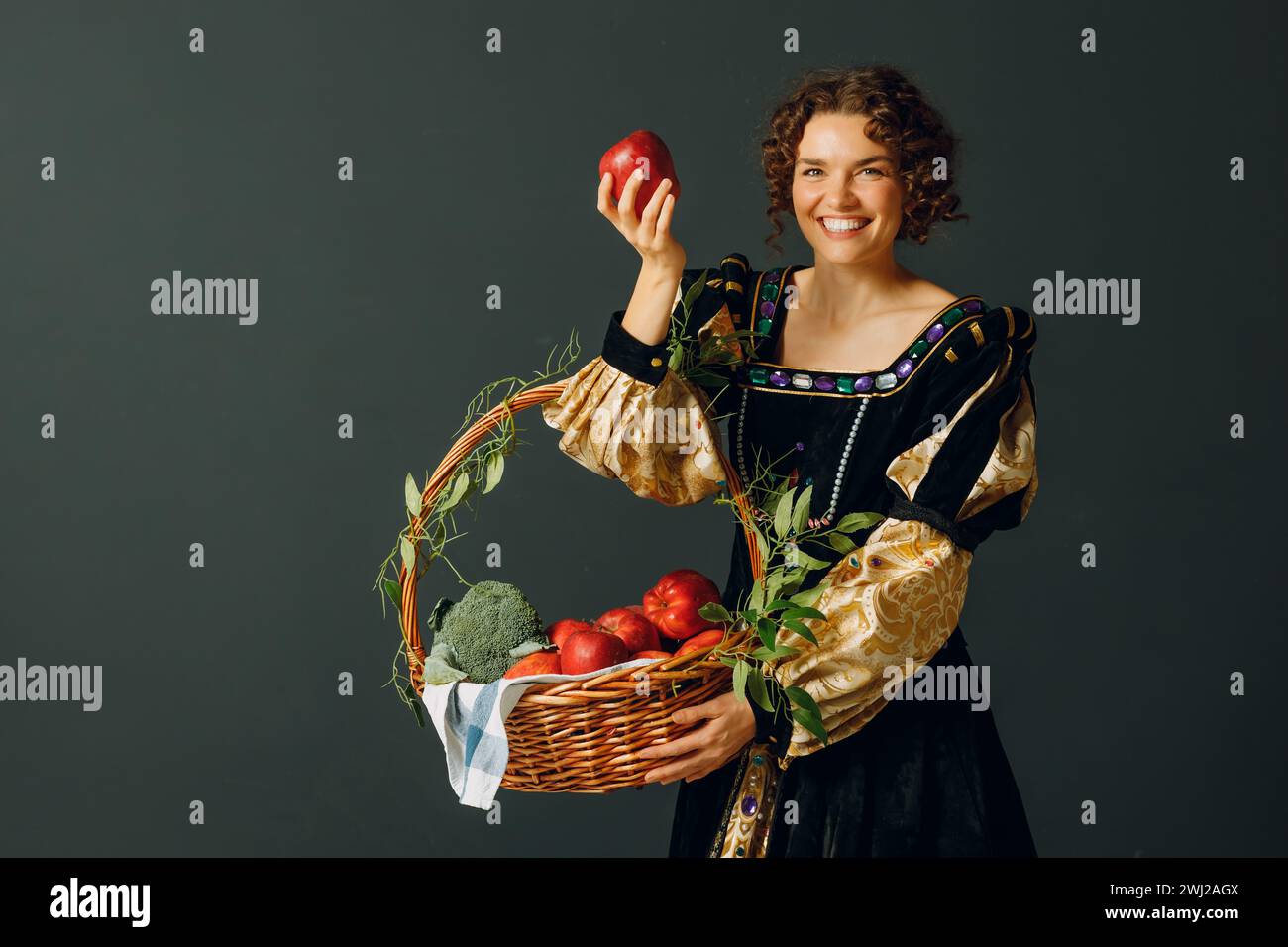 Porträt einer jungen erwachsenen Frau in einem mittelalterlichen Kleid, die einen Korb mit Gemüse und Früchten hält. Ernte und gesunder Foo Stockfoto