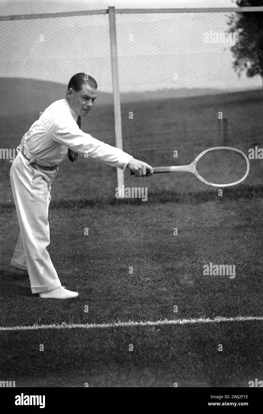 1920er Jahre, historisch, draußen auf einem Rasen-Tennisplatz, ein Gentleman-Tennisspieler in der stilvollen Sportkleidung der Epoche - lange Flanellhose, Langarmshirt und Krawatte - spielt eine Rückhand mit einem traditionellen Holzschläger, England, Großbritannien. Stockfoto