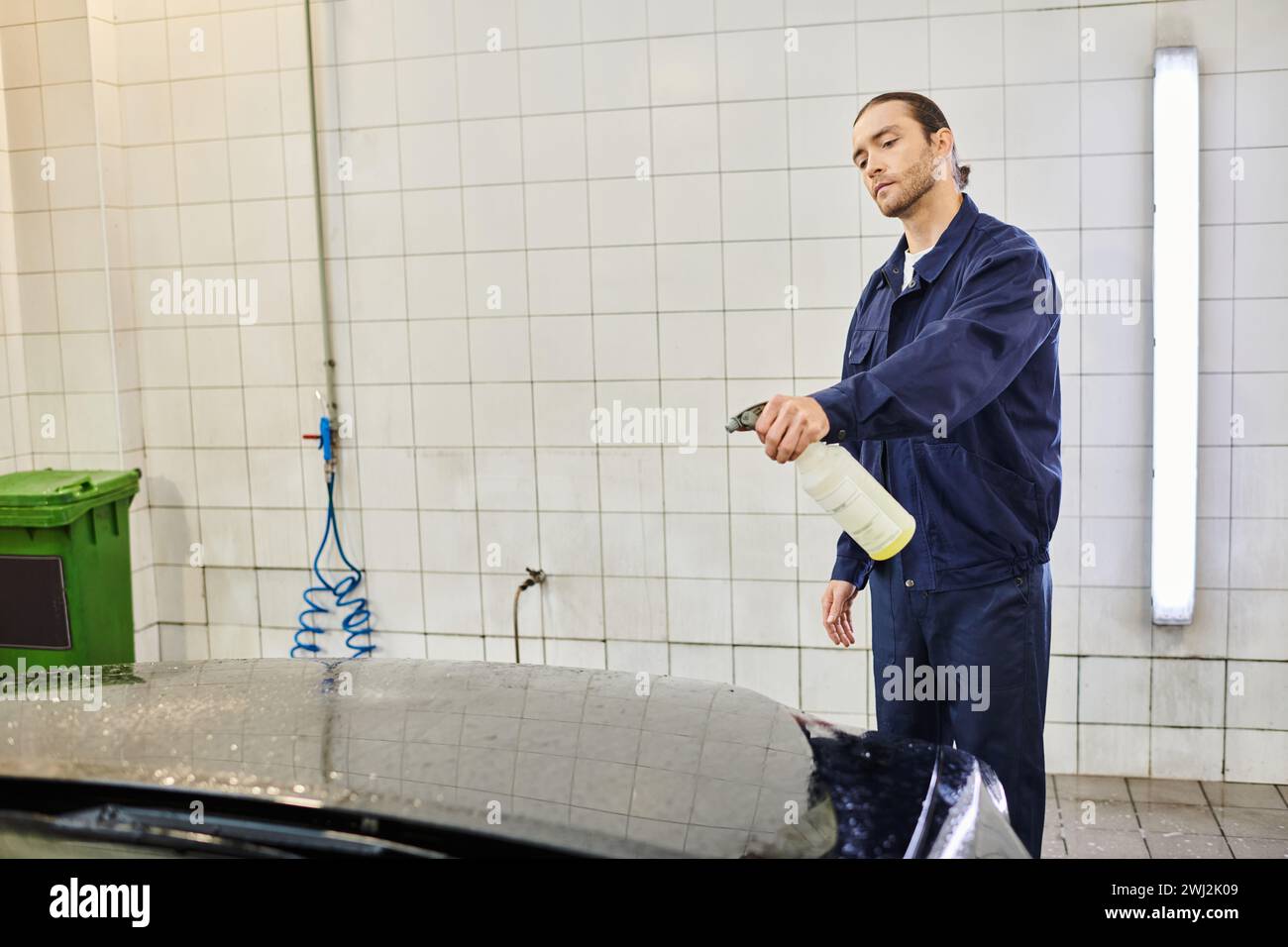 Gut aussehender, hart arbeitender Mann mit gesammelten Haaren in blauer Uniform, mit Pulverisierer, um das Auto zu reinigen Stockfoto