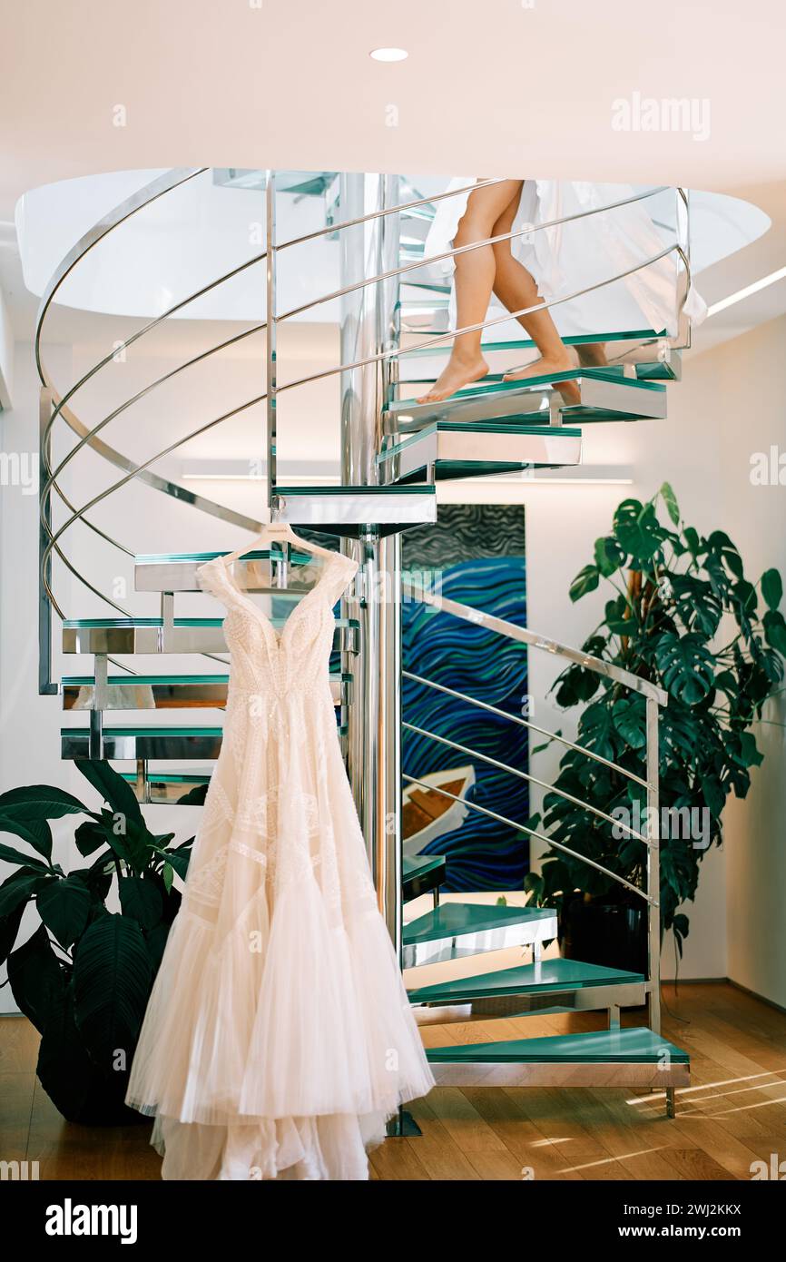 Barfuß Braut im Gewand geht eine Wendeltreppe hinunter zu einem Hochzeitskleid, das an einem Kleiderbügel hängt. Abgeschnitten. Gesichtslos Stockfoto