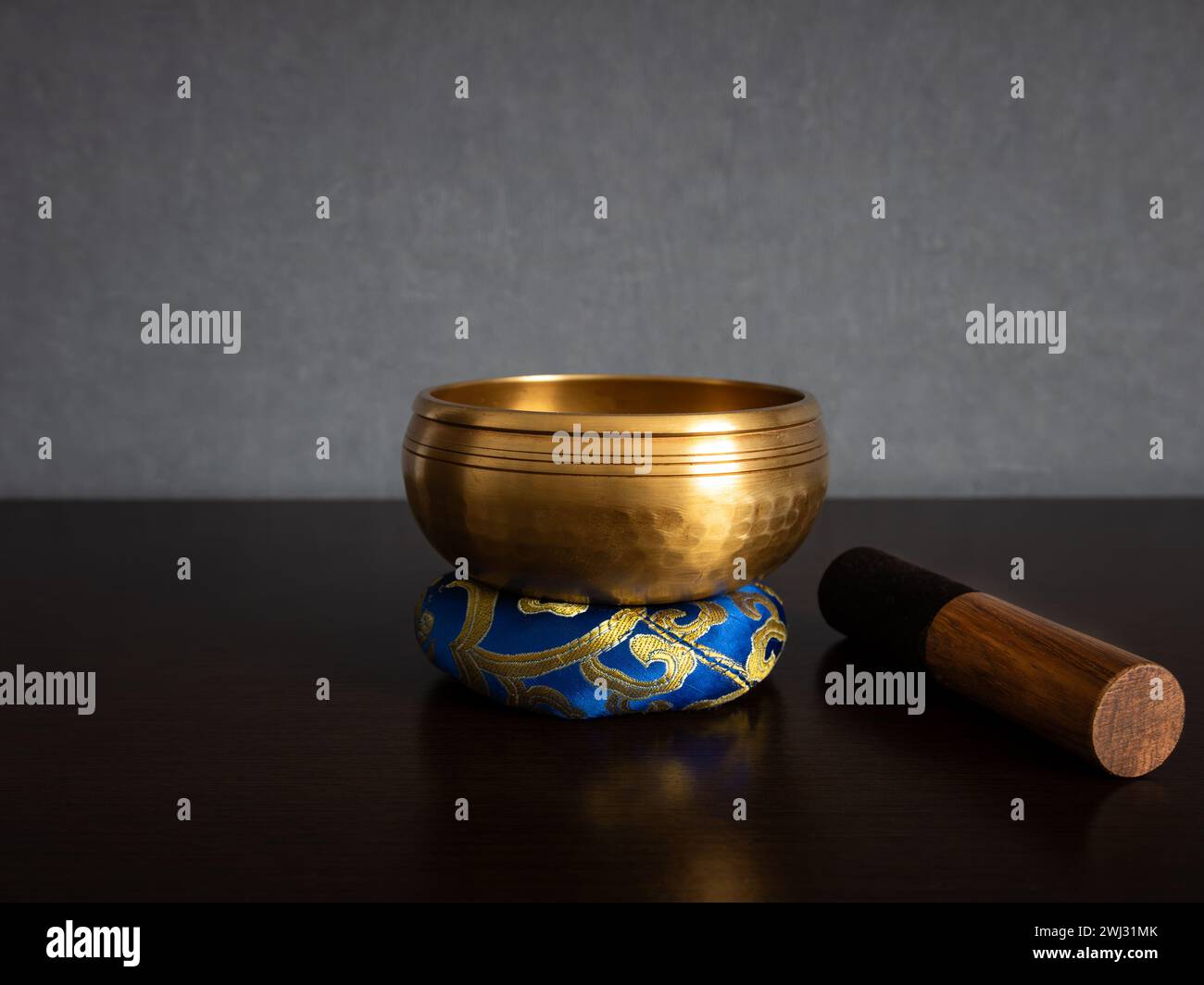 Eine goldfarbene tibetische Klangschale, die auf einem blauen Ringkissen steht, mit einem Streicher an der Seite auf einem unscharfen, sauberen Hintergrund und leerem Platz für Text Stockfoto