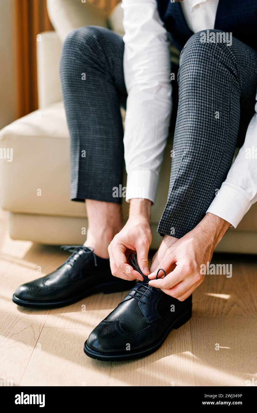 Bräutigam fesselt seine schwarzen Lederbrogues, während er auf dem Sofa sitzt. Abgeschnitten. Gesichtslos Stockfoto