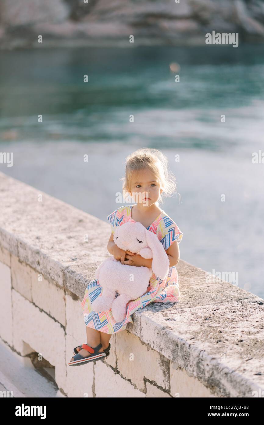 Das kleine Mädchen sitzt auf einem Steinzaun am Meer und umarmt einen Plüschhasen und schaut weg Stockfoto
