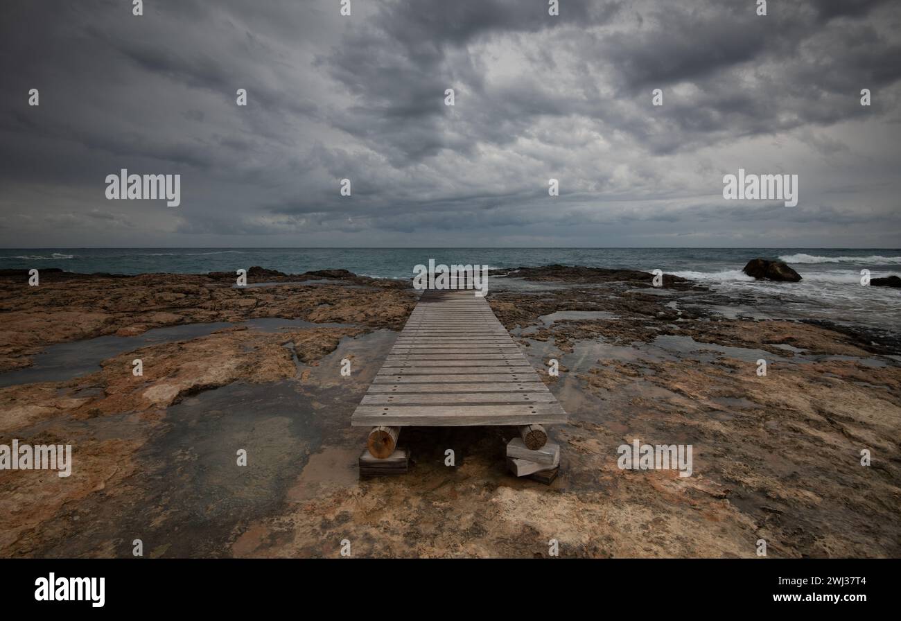 Stürmische Wellen, bewölkter Himmel, hölzerner leerer Pier an einem felsigen Ufer Stockfoto