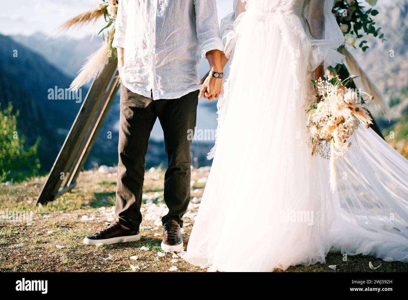 Braut und Bräutigam stehen mit Händen am Hochzeitsbogen auf einem Berg. Abgeschnitten. Gesichtslos Stockfoto