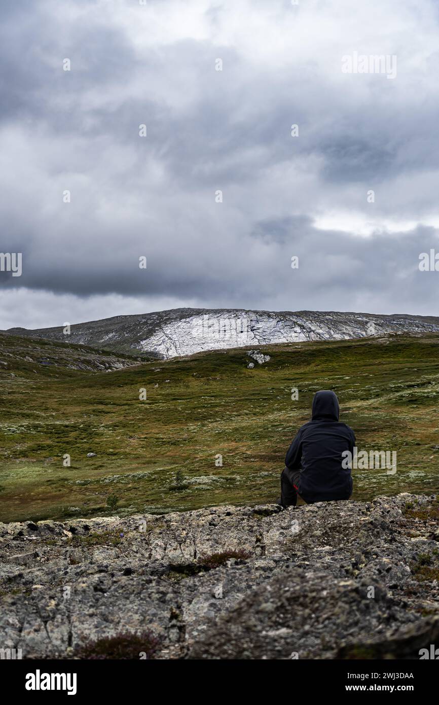 Einsames Kind, das auf felsigem Felsvorsprung sitzt und die weite zerklüftete Landschaft und den weiten Blick auf die weiße Klippe am bewölkten Himmel in Norwegen betrachtet Stockfoto