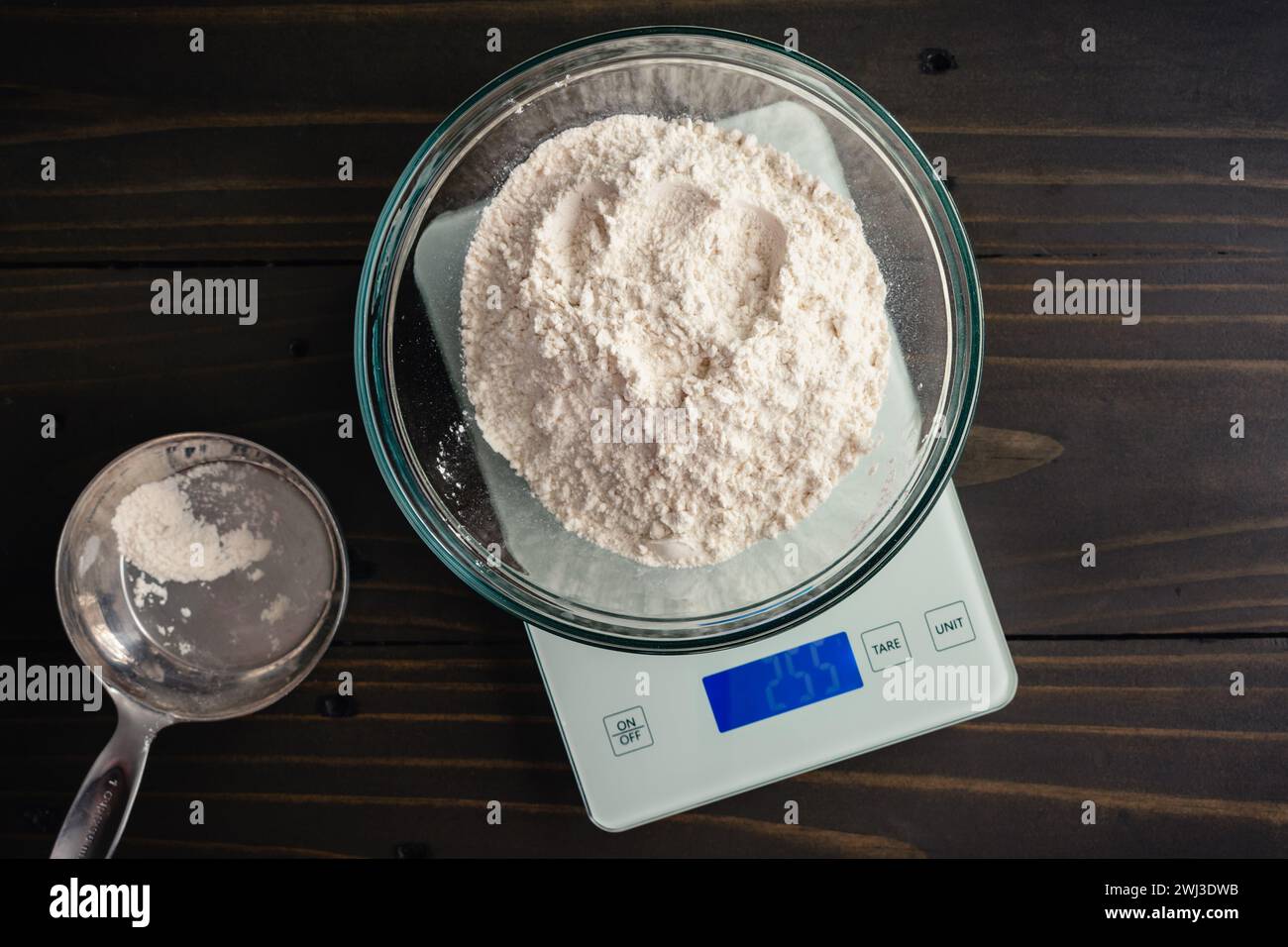 Wiegen einer großen Schüssel mit Allzweckmehl auf einer Küchenwaage: Glasmischschale mit Mehl auf einer digitalen Waage mit einem Messbecher an der Seite Stockfoto