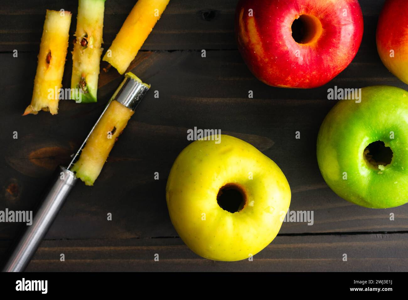 Entkernte Äpfel auf einem rustikalen Holztisch: Ambrosia-, Golden Delicious- und Granny Smith-Äpfel mit einem Entkernungswerkzeug auf dunklem Hintergrund Stockfoto