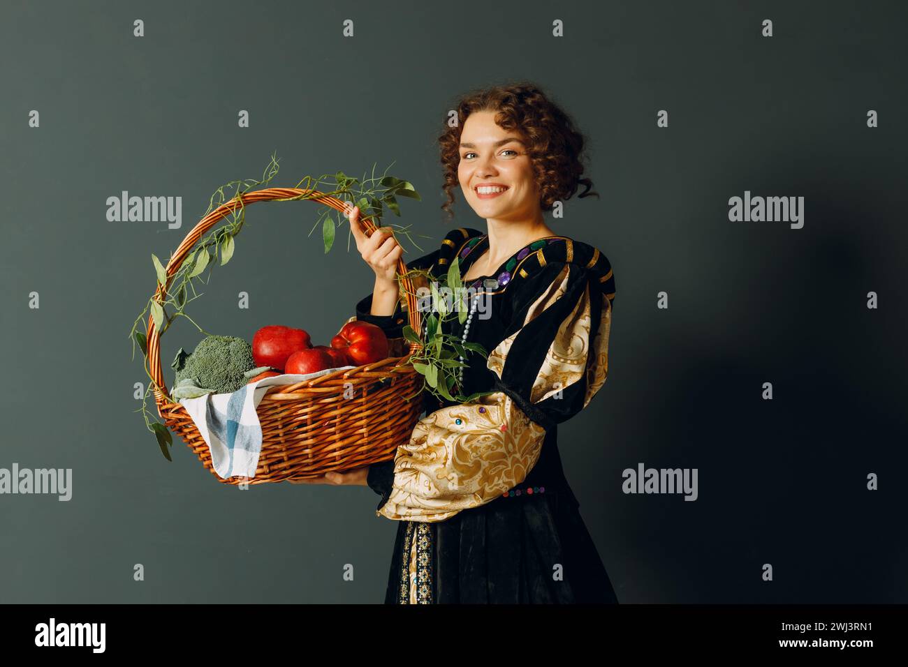 Porträt einer jungen erwachsenen Frau in einem mittelalterlichen Kleid, die einen Korb mit Gemüse und Früchten in der Hand hält. Stockfoto