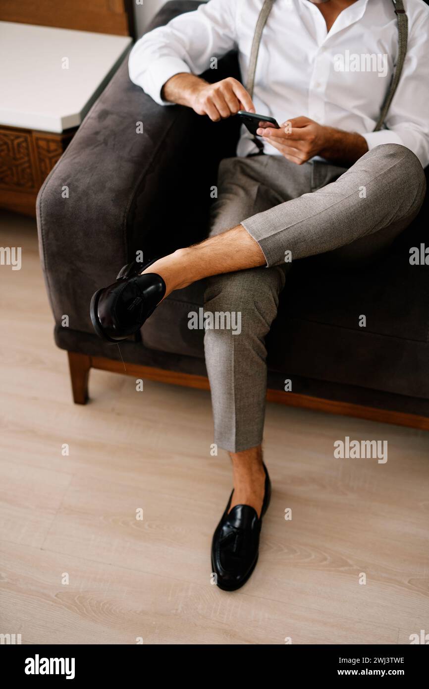 Der Bräutigam klickt auf ein Smartphone in seinen Händen, während er auf dem Sofa sitzt. Abgeschnitten. Gesichtslos Stockfoto