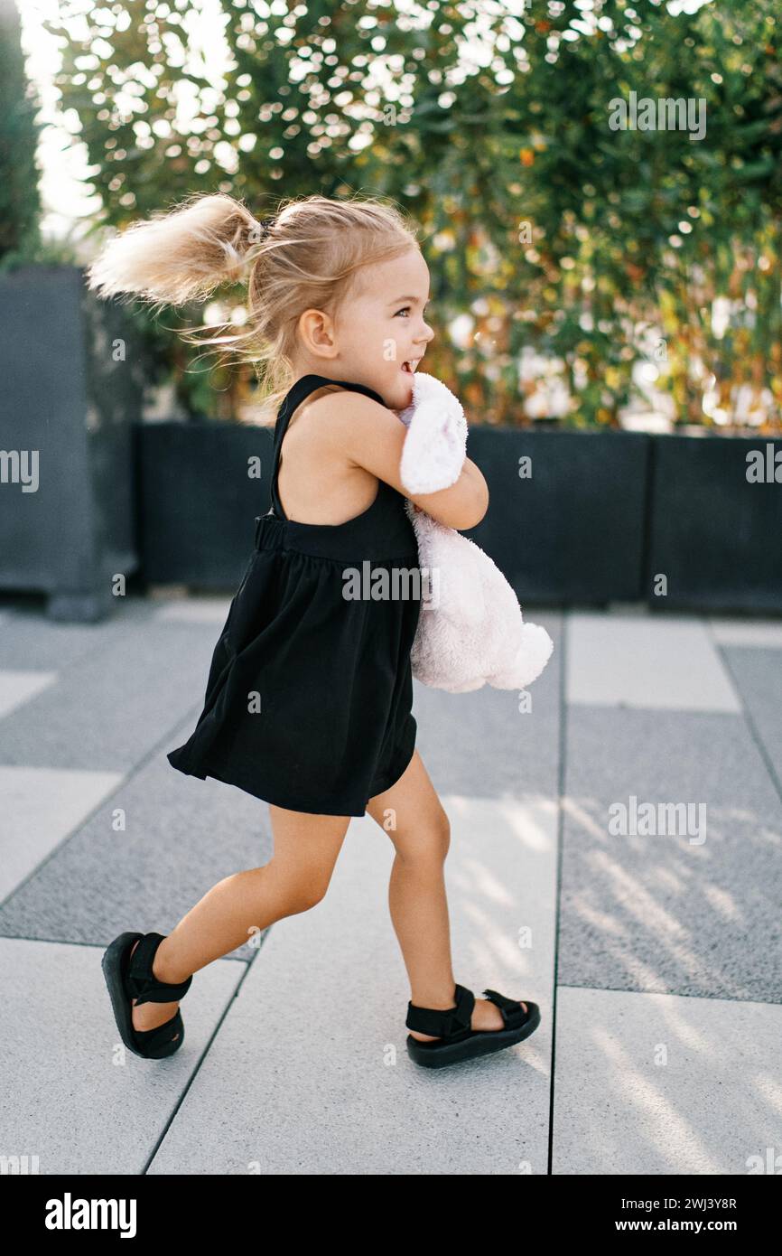 Ein kleines Mädchen mit fliegenden Haaren rennt um den Hof und umarmt einen Spielzeughasen Stockfoto