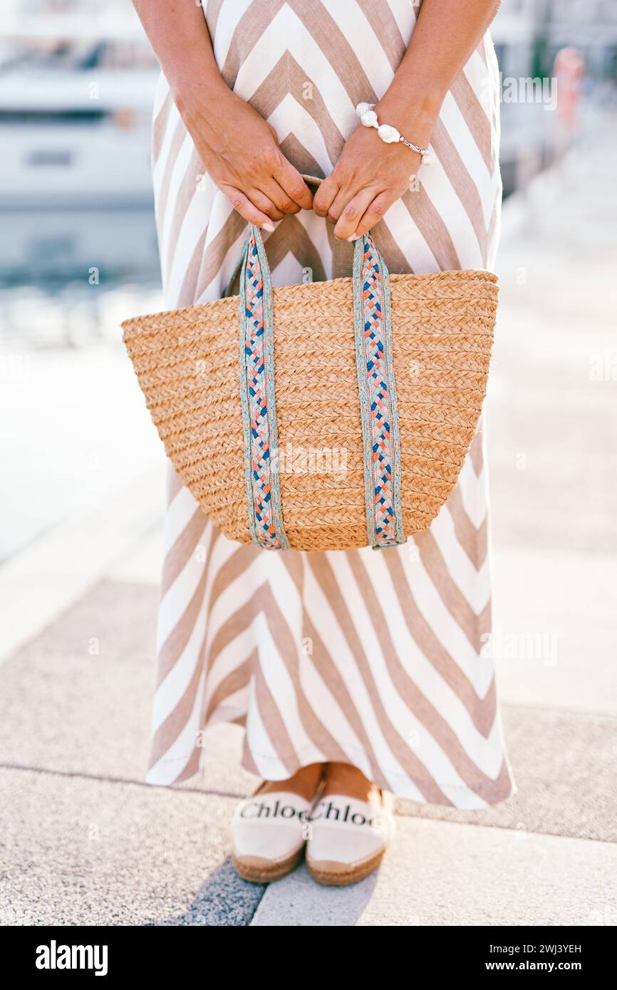 Eine Frau in einem gestreiften Sommerkleid mit einem Strohbeutel in den Händen steht auf dem Pier Stockfoto