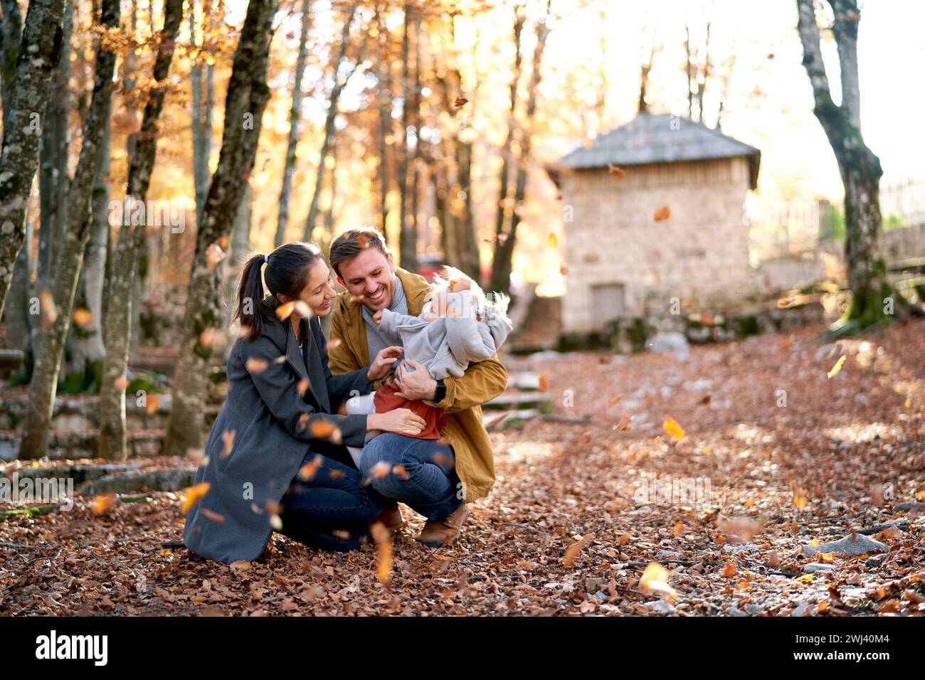 Mom hockt und umarmt Dad mit einem kleinen Mädchen auf den Knien im Park unter fallenden Blättern Stockfoto