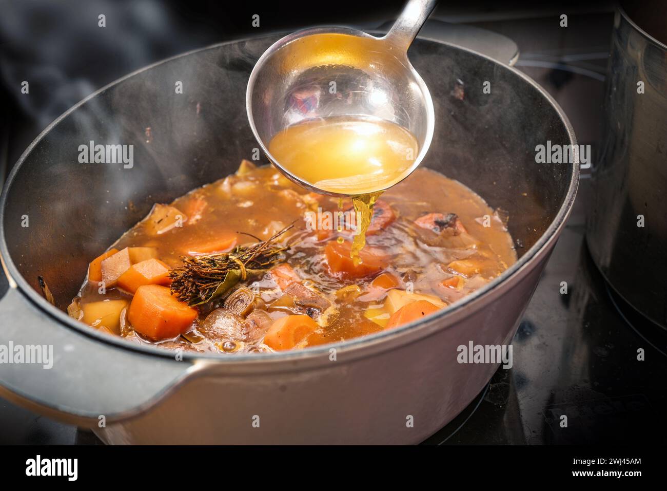 Klare Brühe wird mit einer Kelle in einen Topf mit dampfendem Gemüse wie Karotten, Zwiebeln, Sellerie, Lauch und Kräutern gegossen Stockfoto